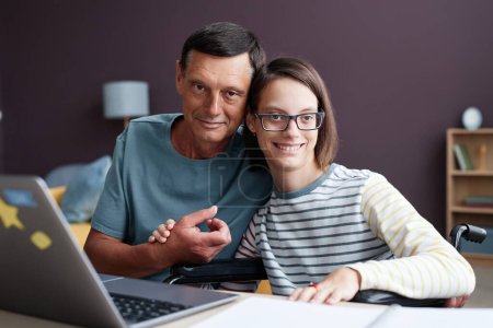 Téléchargez les photos : Portrait d'une adolescente souriante handicapée posant avec son père à la maison et utilisant un ordinateur portable - en image libre de droit