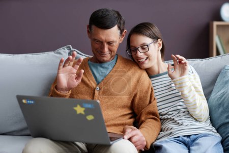 Photo for Portrait of father and teen daughter waving hello to video chat while sitting on sofa with laptop - Royalty Free Image