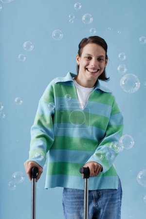 Foto de Retrato vertical de la cintura hacia arriba de una adolescente sonriente con discapacidad mirando la cámara posando con burbujas contra el azul - Imagen libre de derechos