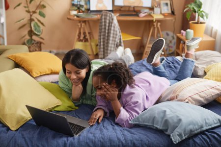 Photo for Portrait of happy black mother and daughter using laptop and watching movie lying on bed together in childrens room - Royalty Free Image