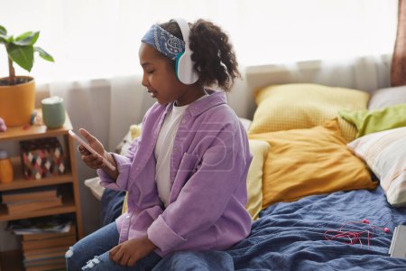 Foto de Retrato de vista lateral de una joven negra usando un teléfono inteligente sentado en la cama y usando auriculares, espacio para copiar - Imagen libre de derechos