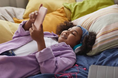Téléchargez les photos : Portrait de fille noire souriante utilisant un smartphone couché sur le lit à la maison et portant un casque - en image libre de droit