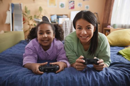Front view portrait of happy mother and daughter playing videogames together lying on bed at home
