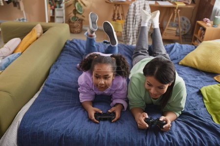 Photo for High angle portrait of happy black mother and daughter playing video games together lying on bed at home - Royalty Free Image