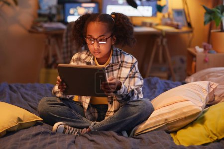 Foto de Retrato de cuerpo entero de adolescente negro con coletas usando tableta digital mientras está sentado en la cama en casa en acogedora luz cálida, espacio de copia - Imagen libre de derechos