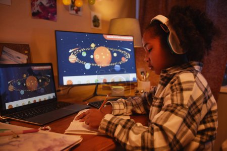 Photo for Side view portrait of teen black girl using computer with space theme while studying at home in cozy evening light, copy space - Royalty Free Image