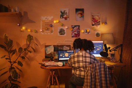 Foto de Vista posterior de la muchacha negra adolescente usando la computadora con el tema del espacio mientras que estudia en el país en la luz de noche acogedora, espacio de la copia - Imagen libre de derechos