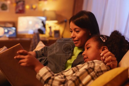 Photo for Portrait of young Black mother and daughter lying in bed together and reading book at night - Royalty Free Image