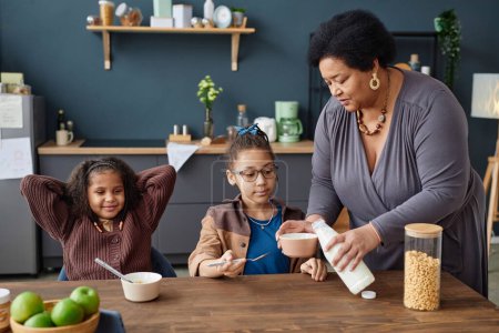 Foto de Retrato de cuidar a la abuela afroamericana que sirve el desayuno a dos niñas en la mesa de la cocina - Imagen libre de derechos