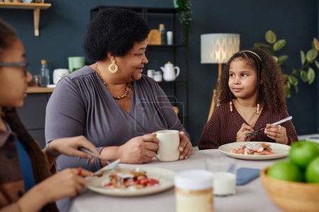 Téléchargez les photos : Portrait de femme noire aînée avec deux enfants dégustant le petit déjeuner ensemble à la table de cuisine - en image libre de droit