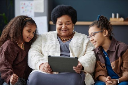Foto de Retrato de vista frontal de una mujer negra mayor sonriente usando una tableta digital en casa con dos chicas jóvenes ayudando - Imagen libre de derechos