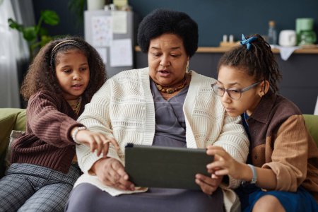 Téléchargez les photos : Portrait avant de la femme âgée noire utilisant une tablette numérique à la maison avec deux enfants aidant - en image libre de droit