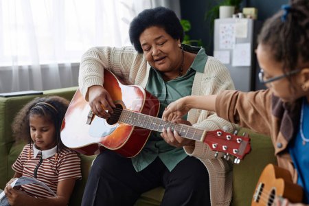 Téléchargez les photos : Portrait d'une femme âgée afro-américaine jouant de la guitare à la maison avec deux petites filles - en image libre de droit