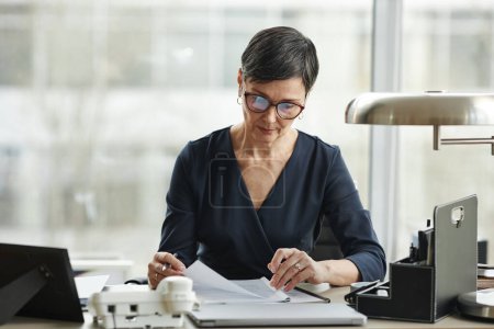 Téléchargez les photos : Portrait frontal d'une élégante femme d'affaires âgée travaillant avec des papiers au bureau dans un bureau privé - en image libre de droit