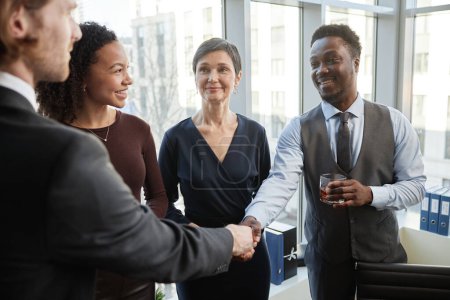 Téléchargez les photos : Groupe diversifié de partenaires d'affaires souriants serrant la main dans le bureau debout avec des boissons et célébrer l'accord - en image libre de droit