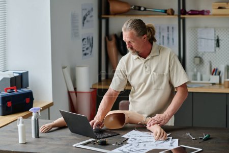 Waist up portrait of long haired senior man making arm prosthetics in workshop and using laptop, copy space