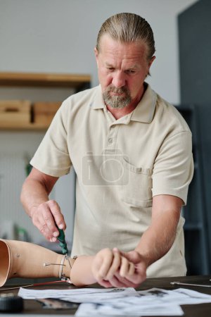 Photo for Vertical portrait of long haired senior man making arm prosthetics in workshop looking at blueprints and schemes - Royalty Free Image