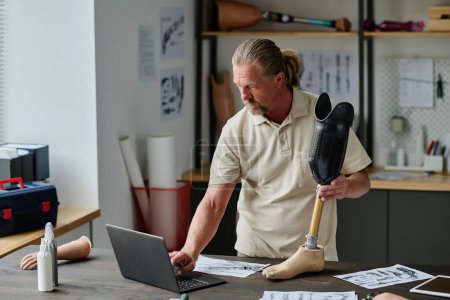 Foto de Retrato de cintura hacia arriba de hombre mayor de pelo largo construyendo prótesis de pierna en el taller y utilizando el ordenador portátil, espacio de copia - Imagen libre de derechos