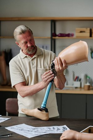 Vertical portrait of long haired senior craftsman building leg prosthetics in workshop and inspecting joints