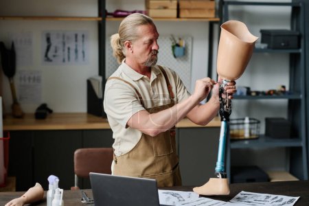 Side view portrait of long haired senior craftsman building handmade leg prosthetics in workshop and screwing knee joint, copy space