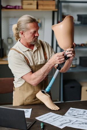 Téléchargez les photos : Portrait vertical d'un homme âgé aux cheveux longs construisant des prothèses de jambes faites à la main dans un atelier d'artisanat et une articulation du genou vissée - en image libre de droit