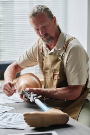 Téléchargez les photos : Portrait vertical de l'artisan senior musclé construisant des prothèses de jambes sur une table en bois avec des schémas et des plans - en image libre de droit