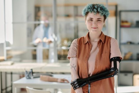 Téléchargez les photos : Portrait taille haute de jeune femme avec un bras prothétique bionique posant dans le bureau des médecins et souriant à la caméra, espace de copie - en image libre de droit