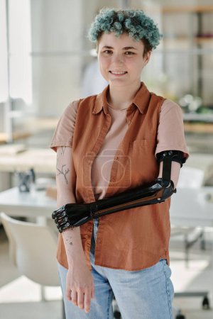 Téléchargez les photos : Portrait vertical de jeune fille avec bras prothétique et cheveux colorés souriant à la caméra dans le bureau des médecins et - en image libre de droit