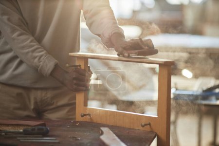 Close up of black man sanding wooden furniture in carpentry workshop lit by sunlight, copy space