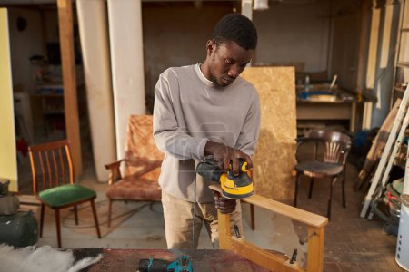 Foto de Retrato tonificado cálido del joven hombre negro construyendo muebles de madera en taller de carpintería y utilizando herramienta de lijado eléctrico, espacio de copia - Imagen libre de derechos