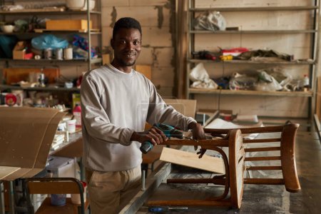 Foto de Retrato de la cintura hacia arriba del hombre afroamericano sonriente restaurando muebles viejos en el taller, espacio de copia - Imagen libre de derechos
