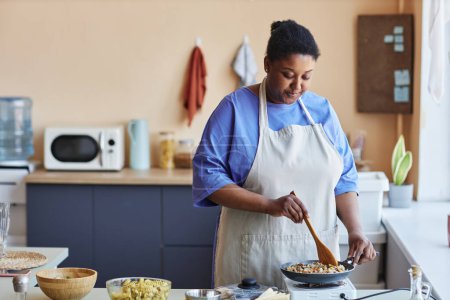 Téléchargez les photos : Portrait taille haute de femme noire souriante frire des légumes et des épices tout en cuisinant dans un espace de copie de cuisine confortable - en image libre de droit