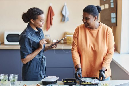Téléchargez les photos : Waist up portrait of two black women reviewing food delivery orders in cafe and using tablet, copy space - en image libre de droit