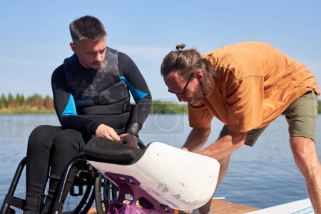 Téléchargez les photos : Portrait d'instructeur sportif senior aidant l'homme handicapé et assemblant du matériel adaptatif pour le wakeboard - en image libre de droit