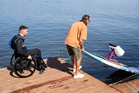 Téléchargez les photos : Portrait latéral d'un instructeur sportif mettant une planche adaptative dans l'eau aidant un homme handicapé à se réveiller - en image libre de droit