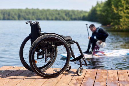 Nahaufnahme eines leeren Rollstuhls von einem hölzernen Steg mit einem behinderten Mann, der im Hintergrund Wassersport genießt, Kopierraum