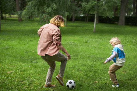 Téléchargez les photos : Action vue arrière du père et du fils jouant au football ensemble dans un parc vert, espace de copie - en image libre de droit