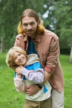 Photo for Vertical portrait of playful father and son playing in park together and embracing looking at camera - Royalty Free Image