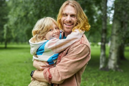 Photo for Waist up portrait of happy father holding blonde little boy in park looking at camera together copy space - Royalty Free Image