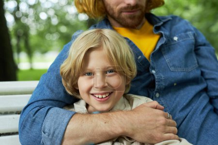 Téléchargez les photos : Gros plan du père et du fils assis sur le banc dans le parc avec le garçon blond souriant à la caméra - en image libre de droit