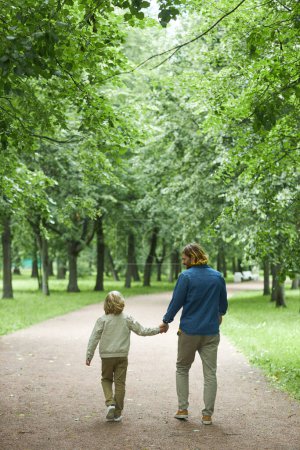 Téléchargez les photos : Vue arrière verticale du père et du fils marchant sur le chemin du parc et tenant la main - en image libre de droit