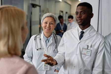 Téléchargez les photos : Jeune médecin généraliste confiant parlant à une assistante tout en se tenant à côté d'un collègue expérimenté heureux lors de la discussion - en image libre de droit