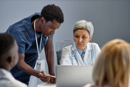 Téléchargez les photos : Jeune assistant confiant en uniforme bleu pointant vers l'écran d'ordinateur portable pendant la présentation et expliquant les données en ligne au médecin expérimenté - en image libre de droit
