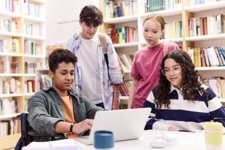 Téléchargez les photos : Groupe multiethnique de jeunes élèves utilisant un ordinateur portable ensemble dans l'intérieur coloré de la bibliothèque à l'école - en image libre de droit