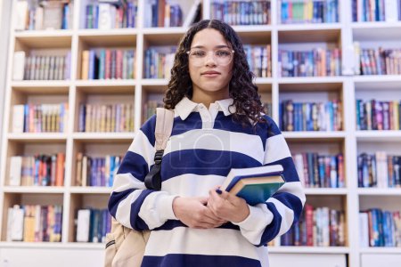 Foto de Retrato de cintura hacia arriba de una joven adolescente con gafas de pie en la biblioteca de la escuela y sosteniendo libros con espacio para copiar la mochila - Imagen libre de derechos