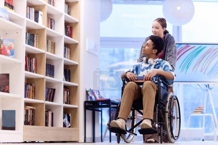 Foto de Retrato de cuerpo entero del adolescente con discapacidad eligiendo libros en la biblioteca de la escuela con una chica sonriente ayudando a copiar espacio - Imagen libre de derechos