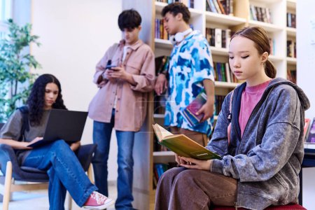 Téléchargez les photos : Vue latérale portrait d'une adolescente asiatique lisant un livre dans le salon de la bibliothèque de l'école avec un groupe d'étudiants copier l'espace - en image libre de droit