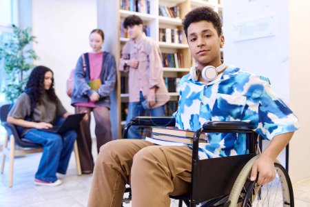 Foto de Retrato de adolescente en silla de ruedas y mirando a la cámara en el moderno salón de la escuela con los estudiantes en el espacio de copia de fondo - Imagen libre de derechos