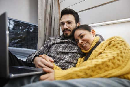 Téléchargez les photos : Portrait de jeune couple souriant en van remorque utilisant l'ordinateur sur le lit ensemble tout en appréciant le voyage confortable en hiver - en image libre de droit