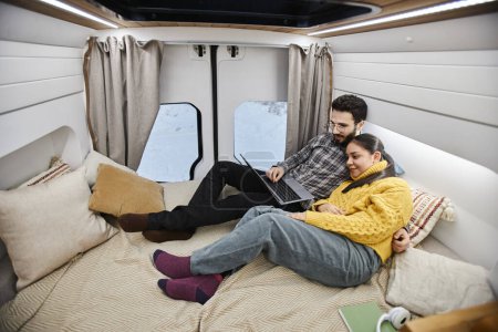 Téléchargez les photos : Portrait en grand angle d'un jeune couple souriant dans une caravane allongée sur le lit ensemble et utilisant un ordinateur tout en profitant d'un voyage confortable en hiver - en image libre de droit